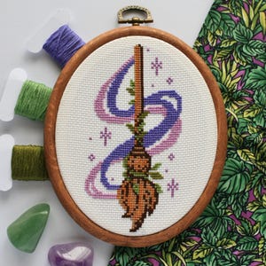 Puede incluir: Un bordado de punto de cruz en un aro de madera presenta un diseño de escoba con cintas moradas y rosas. La escoba es marrón y naranja, con hojas verdes. Bobinas de hilo morado y verde y piedras preciosas rodean el aro.