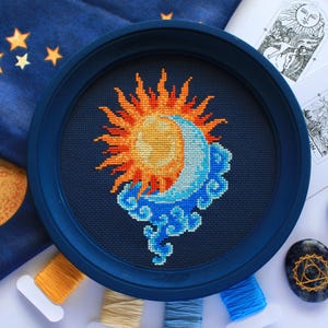 以下が含まれることがあります： 紺色の刺繍枠に収められた、暗い布地に太陽と月が描かれた円形のクロスステッチ刺繍。太陽はオレンジと黄色の光線を放ち、月は水色で、様式化された青い雲の上に配置されています。刺繍は中央に配置されています。