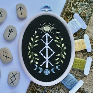 Puede incluir: Bastidor de bordado de punto de cruz ovalado con un diseño blanco y verde claro sobre fondo negro. El diseño incluye un sol, estrellas, un símbolo central y medias lunas. Las runas están dispersas alrededor del bastidor, con carretes de hilo en la esquina.