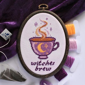Op de afbeelding: Ovale kruissteekborduurwerk in een donkerhouten frame. Het ontwerp toont een paarse beker met een maan en sterren, stoom en de woorden "witches brew". Spoelen garen in oranje, roze en paars zijn zichtbaar.