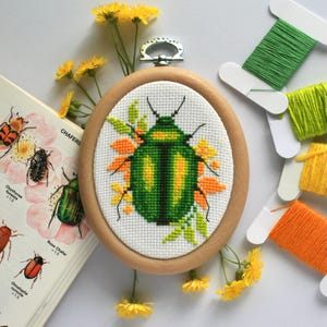 Op de afbeelding: Ovale kruissteekborduurwerk van een groene en gele kever, gezet in een houten hoepel. Het ontwerp is omgeven door oranje en gele bloemaccenten. Een vintage boek met insectenillustraties en spoelen met kleurrijk garen zijn in de buurt.