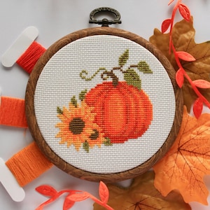 May include: A cross-stitch embroidery hoop featuring an orange pumpkin and sunflower design. The embroidery is set in a wooden hoop with a metal clasp. Orange thread spools and fall leaves surround the hoop.