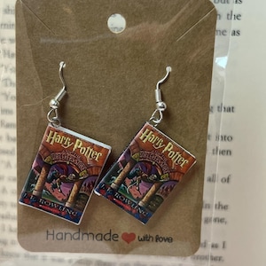 Op de afbeelding: Zilverkleurige oorhangers met miniatuur boekcover designs van "Harry Potter en de Steen der Wijzen" van J.K. Rowling. De oorbellen worden getoond op een bruine kaart met de tekst "Handmade with love".