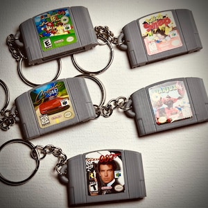 May include: A collection of gray Nintendo 64 game cartridge keychains. Each keychain features a different game label, including "Super Mario 64", "Pokemon Snap", "Star Fox 64", "Cruis'n USA", and "007".