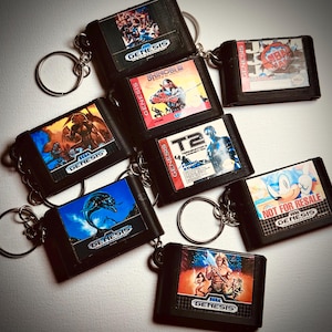 Op de afbeelding: Een verzameling zwarte sleutelhangers met miniatuur Sega Genesis game cartridges. Elke cartridge toont kleurrijke game-afbeeldingen en titels zoals "Shinobi" en "Sonic". De sleutelhangers zijn gerangschikt op een licht oppervlak, met een retro gaming thema.