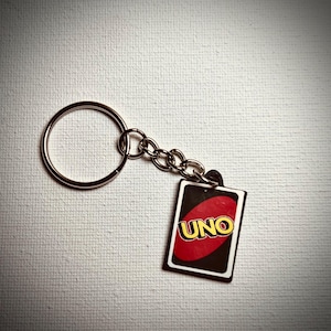 May include: A keychain featuring a small UNO card. The card has a black background with a red circle and the word "UNO" in yellow. The keychain has a silver-colored ring and chain.