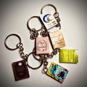 May include: Keychains featuring miniature book replicas. One keychain displays "Alice in Wonderland" with a floral wreath. Other books include "The Secret History" and "Hooky". Each book is attached to a silver chain and ring.
