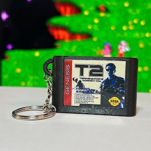 May include: Black keychain featuring a miniature replica of a Sega Genesis video game cartridge. The cartridge has the "T2: Terminator 2: Judgment Day" game label. The keychain includes a silver-colored split ring and chain.