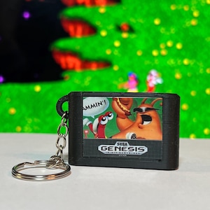 May include: A black Sega Genesis game cartridge keychain. The cartridge features a colorful graphic with the text "AMMIN'!" and the Sega Genesis logo. The keychain has a silver metal ring.