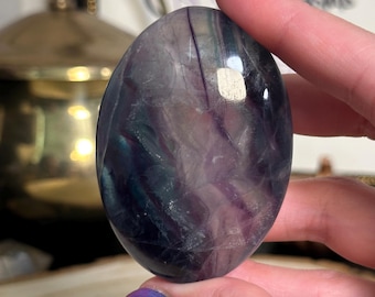 Puede incluir: Una piedra de fluorita pulida, de forma ovalada, con bandas moradas, verdes y claras. La piedra se sostiene en una mano, mostrando su superficie lisa y brillante. El fondo incluye un objeto dorado y una imagen enmarcada.