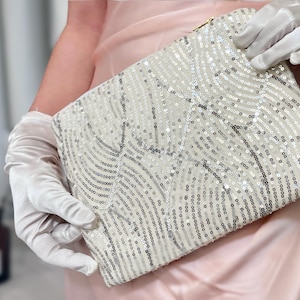 Puede incluir: Clutch rectangular blanco roto cubierta de lentejuelas brillantes dispuestas en un patrón ondulado. El bolso de mano tiene una cremallera dorada y es sostenido por una persona con guantes blancos.