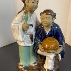 May include: A ceramic figurine depicting two figures in traditional attire. One figure stands, holding a green and gold object, while the other kneels, holding a brown bowl. The figurine features a brown base and is painted with blue, white, and green accents.