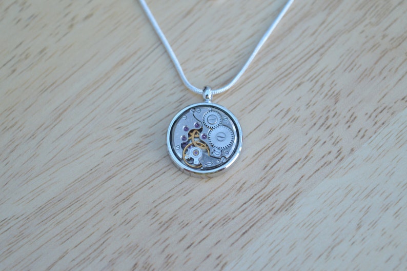 Op de afbeelding: Zilveren ketting met een ronde hanger met een zichtbaar uurwerkmechanisme. De hanger heeft een zilveren afwerking en is versierd met kleine paarse juweeltjes.