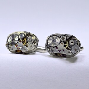 May include: A pair of silver cufflinks made from vintage watch parts. The cufflinks feature intricate gears and a circular face with a silver dial.