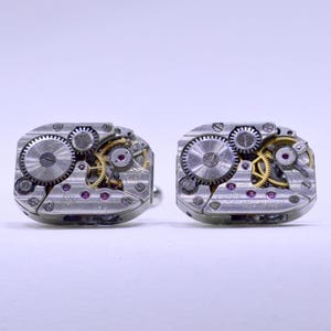 Può includere: Un paio di gemelli argentati che presentano gli intricati meccanismi interni di un orologio. I gemelli rettangolari mostrano ingranaggi, viti e gioielli in un design meccanico dettagliato. Gli ingranaggi sono argentati e dorati, con piccoli gioielli rossi.