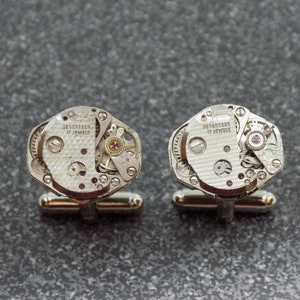 Può includere: Un paio di gemelli d'argento con il meccanismo interno di un movimento di orologio esposto. Ogni gemello ha una base d'argento e un quadrante dell'orologio circolare con il testo "SEVENTEEN 17 JEWELS".