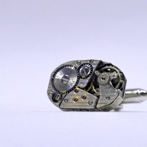 Pode incluir: Abotoaduras de prata com um mecanismo de relojoaria detalhado e exposto. As abotoaduras têm um acabamento prateado e apresentam um design complexo de engrenagens e molas.