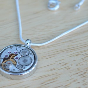 Op de afbeelding: Een zilveren ketting met een ronde hanger met een zichtbaar uurwerkmechanisme met kleine roze juweeltjes.