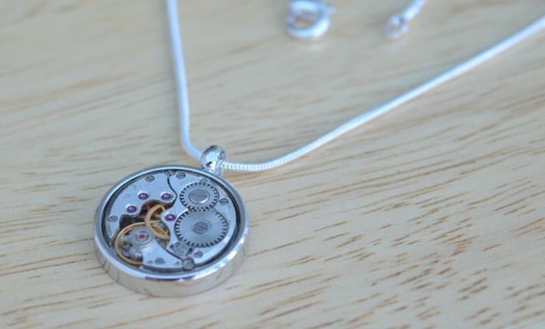 Op de afbeelding: Een zilveren ketting met een ronde hanger met een zichtbaar uurwerkmechanisme met kleine roze juweeltjes.