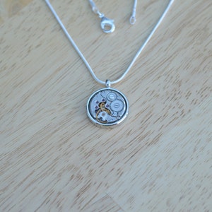Op de afbeelding: Een zilveren ketting met een ronde hanger met een gedetailleerd uurwerkmechanisme. De hanger is zilverkleurig met een helder glazen oppervlak dat de innerlijke werking van een horloge onthult.