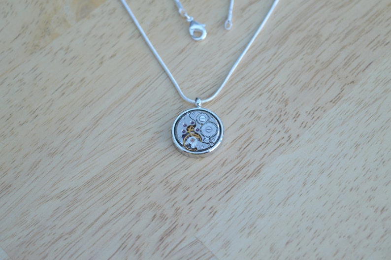 Op de afbeelding: Een zilveren ketting met een ronde hanger met een gedetailleerd uurwerkmechanisme. De hanger is zilverkleurig met een helder glazen oppervlak dat de innerlijke werking van een horloge onthult.
