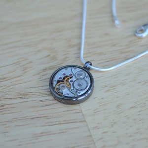 Op de afbeelding: Een zilveren ketting met een ronde hanger met een zilveren en gouden uurwerk.