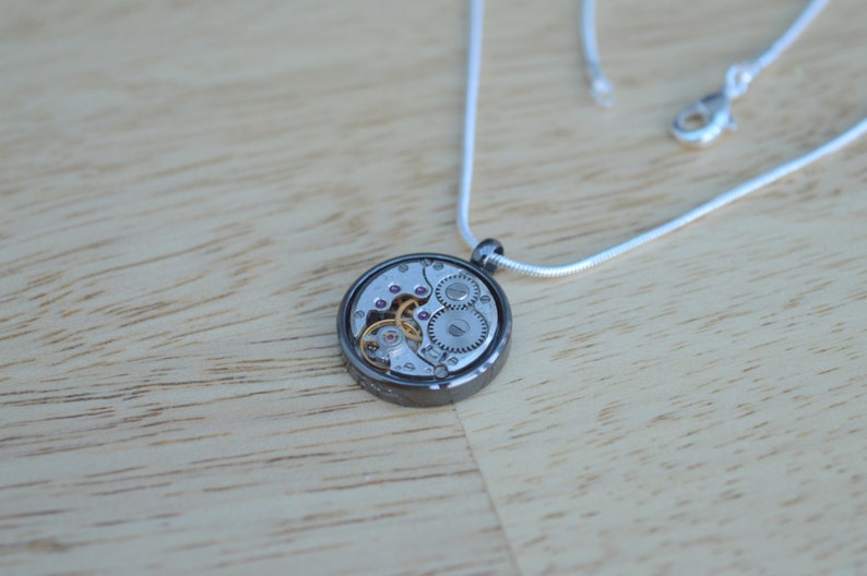 Op de afbeelding: Een zilveren ketting met een ronde hanger met een zilveren en gouden uurwerk.