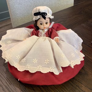 Puede incluir: Una muñeca vintage con cabello castaño, vestida con un vestido rojo y un gorro blanco con un lazo negro. El vestido de la muñeca tiene un cuello de encaje blanco y está rodeado por una falda blanca con bordados florales. La muñeca está sentada sobre un cojín rojo.