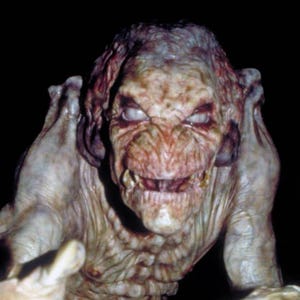Puede incluir: Una criatura grotesca de piel pálida con rostro arrugado, dientes afilados y ojos blancos brillantes. El monstruo tiene orejas grandes y extiende sus manos con garras, sobre un fondo oscuro. El tema general es el terror.