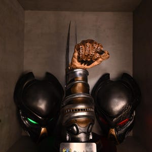 Könnte beinhalten: Eine Präsentation von Predator-Sammlerstücken. Das zentrale Stück ist eine detaillierte Skulptur eines Predator-Handschuhs mit einer Klinge. Flankiert wird es von zwei Predator-Masken, eine mit grünen und die andere mit roten leuchtenden Augen. Die Basis trägt den Text "DoubleG".