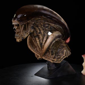 May include: A detailed sculpture of an Alien Xenomorph head and upper body, with a glossy, dark brown head and a textured, tan body. The creature is mounted on a black base, with a partially eaten apple nearby. The background is black.