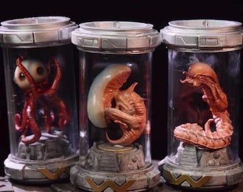 Könnte beinhalten: Drei zylindrische Glasbehälter mit silbernen Deckeln, die jeweils eine andere außerirdische Kreatur beherbergen. Einer hat eine rote, oktopusartige Kreatur, ein anderer einen beigen Embryo und der dritte einen rötlich-orangen Alien. Die Behälter stehen auf einem dekorativen Sockel.