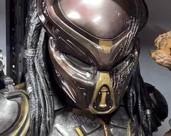 May include: A detailed bust of the Predator alien, featuring a metallic silver, bronze, and gold color scheme. The bust includes the Predator's iconic helmet, dreadlocks, and armor, with the word "Predator" on a small plaque.
