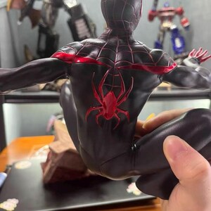 May include: A black action figure of a character in a suit with a red spider emblem on the back and red web patterns across the shoulders and head. The figure is posed in a crouched position with arms extended outward, showcasing detailed textures.