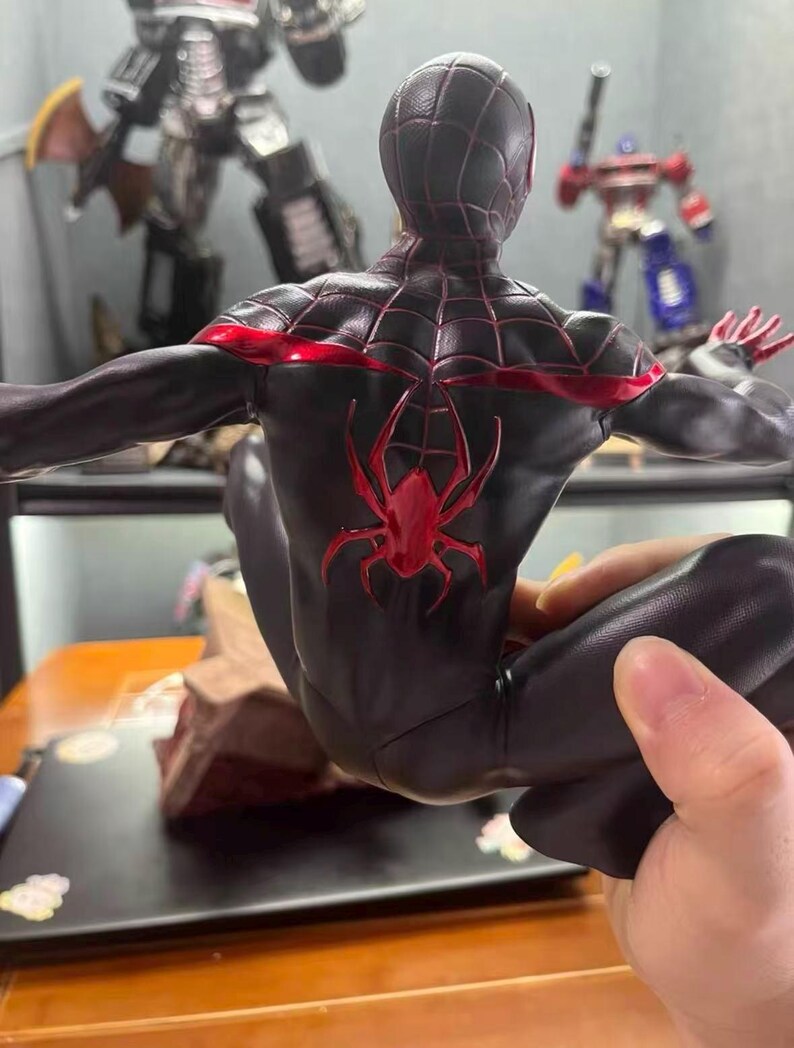 May include: A black action figure of a character in a suit with a red spider emblem on the back and red web patterns across the shoulders and head. The figure is posed in a crouched position with arms extended outward, showcasing detailed textures.