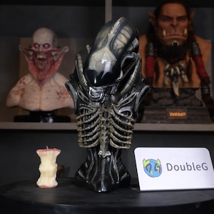 May include: A black and beige Xenomorph bust featuring a smooth elongated head, exposed ribcage structure, and intricate mechanical tubing details. Beside it sits a small apple core candle and a white sign with the text DoubleG in blue and green.