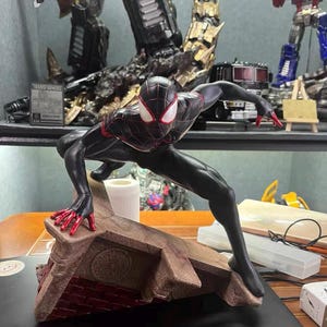 May include: A Miles Morales Spider-Man action figure in a dynamic crouching pose. The figure is black with red web patterns, red fingertips, and white eye lenses. It is mounted on a textured, brown, triangular base resembling a brick rooftop.