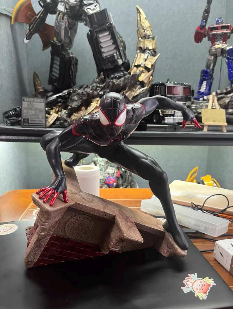 May include: A Miles Morales Spider-Man action figure in a dynamic crouching pose. The figure is black with red web patterns, red fingertips, and white eye lenses. It is mounted on a textured, brown, triangular base resembling a brick rooftop.