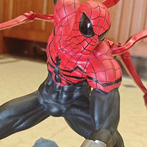 May include: A detailed figurine of Spider-Man in a dynamic pose. The figure features a red and black costume with web detailing, black eye coverings, and metallic red appendages extending from the back. The figure is on a base with a gray rock-like structure.
