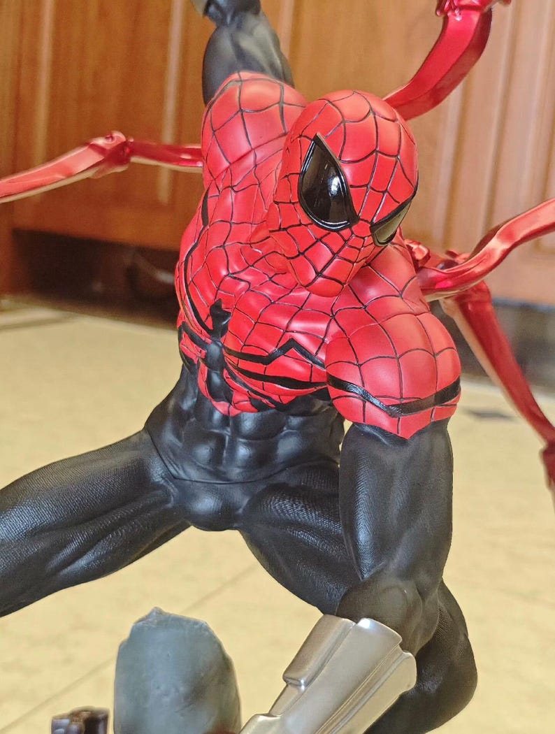 May include: A detailed figurine of Spider-Man in a dynamic pose. The figure features a red and black costume with web detailing, black eye coverings, and metallic red appendages extending from the back. The figure is on a base with a gray rock-like structure.