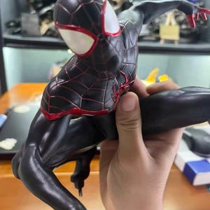 May include: A black and red action figure of a superhero character in a dynamic crouching pose. The figure features a textured black suit with red web patterns, a red spider emblem on the chest, and large white eye lenses on the mask.