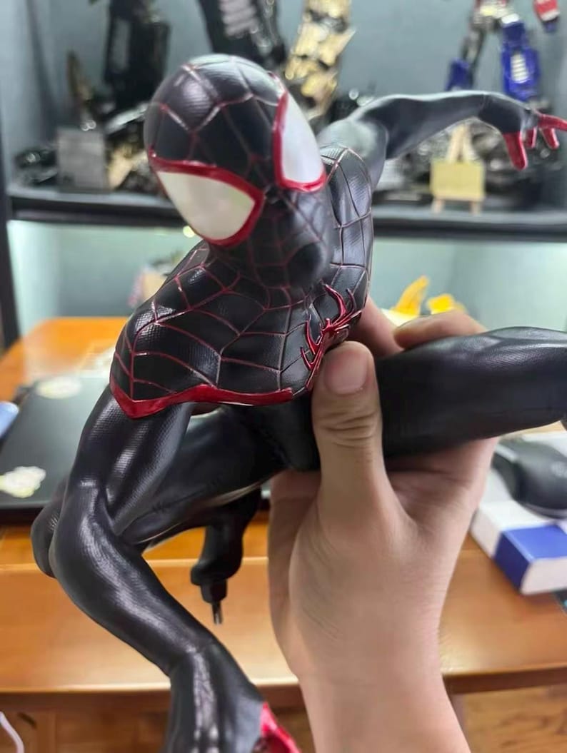 May include: A black and red action figure of a superhero character in a dynamic crouching pose. The figure features a textured black suit with red web patterns, a red spider emblem on the chest, and large white eye lenses on the mask.