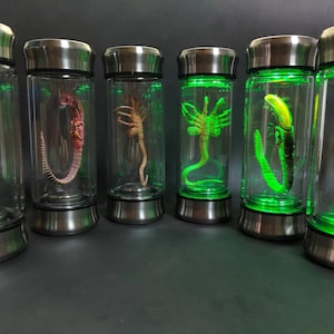 May include: Six cylindrical glass display canisters with metallic silver caps and bases. Four of the canisters contain detailed creature figurines, while two are empty. The canisters are illuminated from the base with a vibrant green light.
