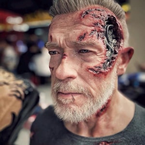May include: A close-up of a man's face with special effects makeup. The makeup simulates a cyborg with exposed mechanical components and realistic wounds. The man has gray hair and a gray beard, wearing a dark gray shirt.