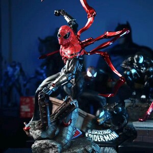 May include: A detailed action figure of Spider-Man in a dynamic pose, featuring a red and black suit with metallic red spider-arms. The figure is set on a rocky base with the words "Amazing Spider-Man" visible. Other superhero figures are in the background.