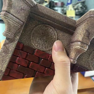 May include: A triangular display base featuring a textured brown stone-like finish with a central circular emblem and a section of dark red brickwork. The object has raised corner supports and a weathered appearance with visible surface detailing.