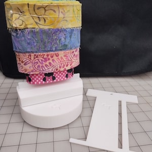 May include: A white plastic ribbon storage organizer with five spools of ribbon. The ribbons are in various patterns and colors, including yellow, blue, pink, and white. The organizer has a base and a vertical stand with slots for the ribbon spools.
