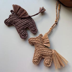 Puede incluir: Dos adornos de caballo hechos a mano con ganchillo. Uno es de color marrón oscuro y el otro de color marrón claro. Ambos tienen crin y cola de rafia. Uno tiene una cuerda con cuentas de madera.