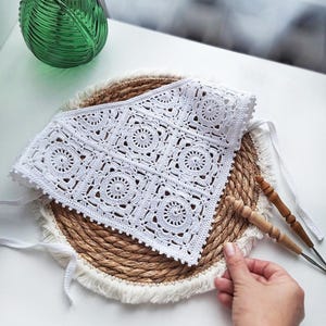 Könnte beinhalten: Ein quadratischer, weißer, gehäkelter Artikel mit floralem Muster, der auf einem runden, gewebten Platzset mit Fransenrand liegt. Der Artikel hat ein zartes, durchbrochenes Design und ist mit weißen Schnüren gebunden. Eine grüne Glasvase mit weißen Blumen befindet sich im Hintergrund.