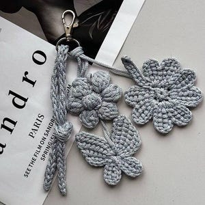 Puede incluir: Un llavero de ganchillo gris con tres colgantes florales y de hojas. El llavero incluye una borla trenzada y un cierre de metal. Los colgantes incluyen una flor, un trébol de cuatro hojas y una hoja.